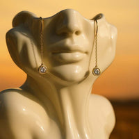 Elegante goldene Threader-Ohrringe mit funkelnden Zirkonia, fotografiert bei Sonnenuntergang, präsentiert auf einer abstrakten Keramik-Büste – DKeniz Schmuck Online-Shop Nürnberg, Bayern.