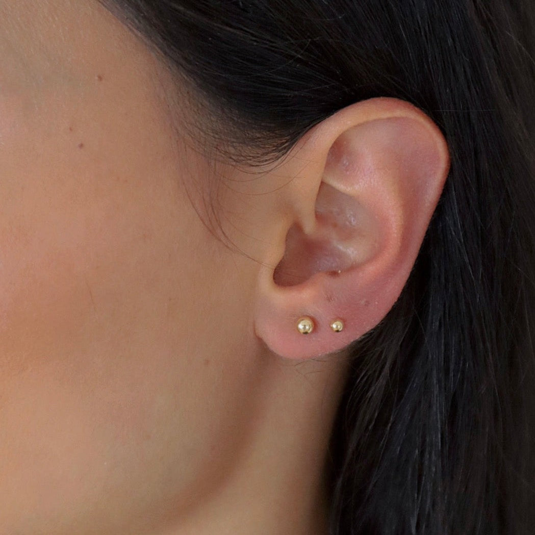 Zwei goldene Balls M Stud Ohrstecker in unterschiedlichen Größen dezent am Ohr einer Frau getragen.