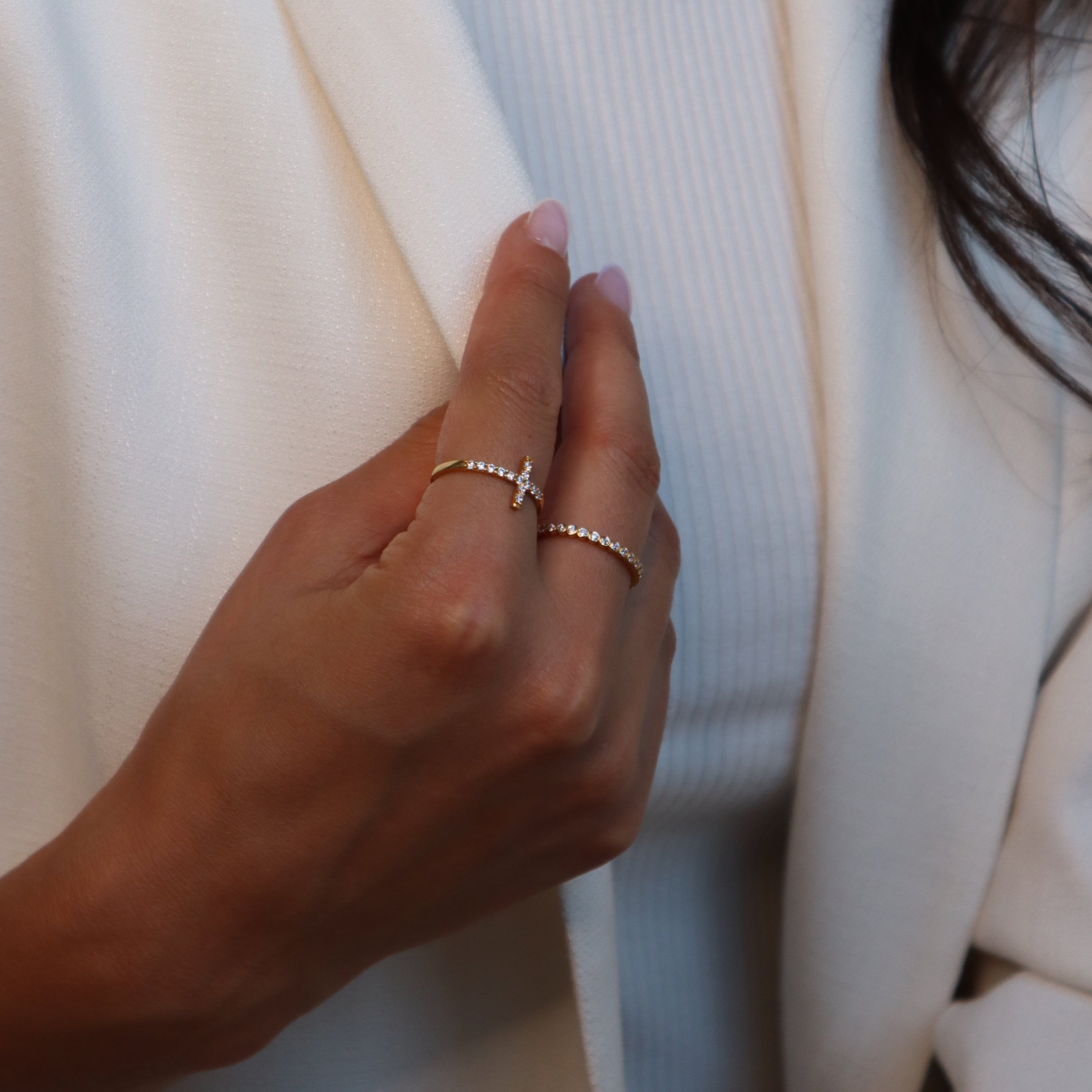 Hand mit Cross Ring in Gold und zartem Ring daneben, getragen zu weißem Blazer, filigraner Damenring.