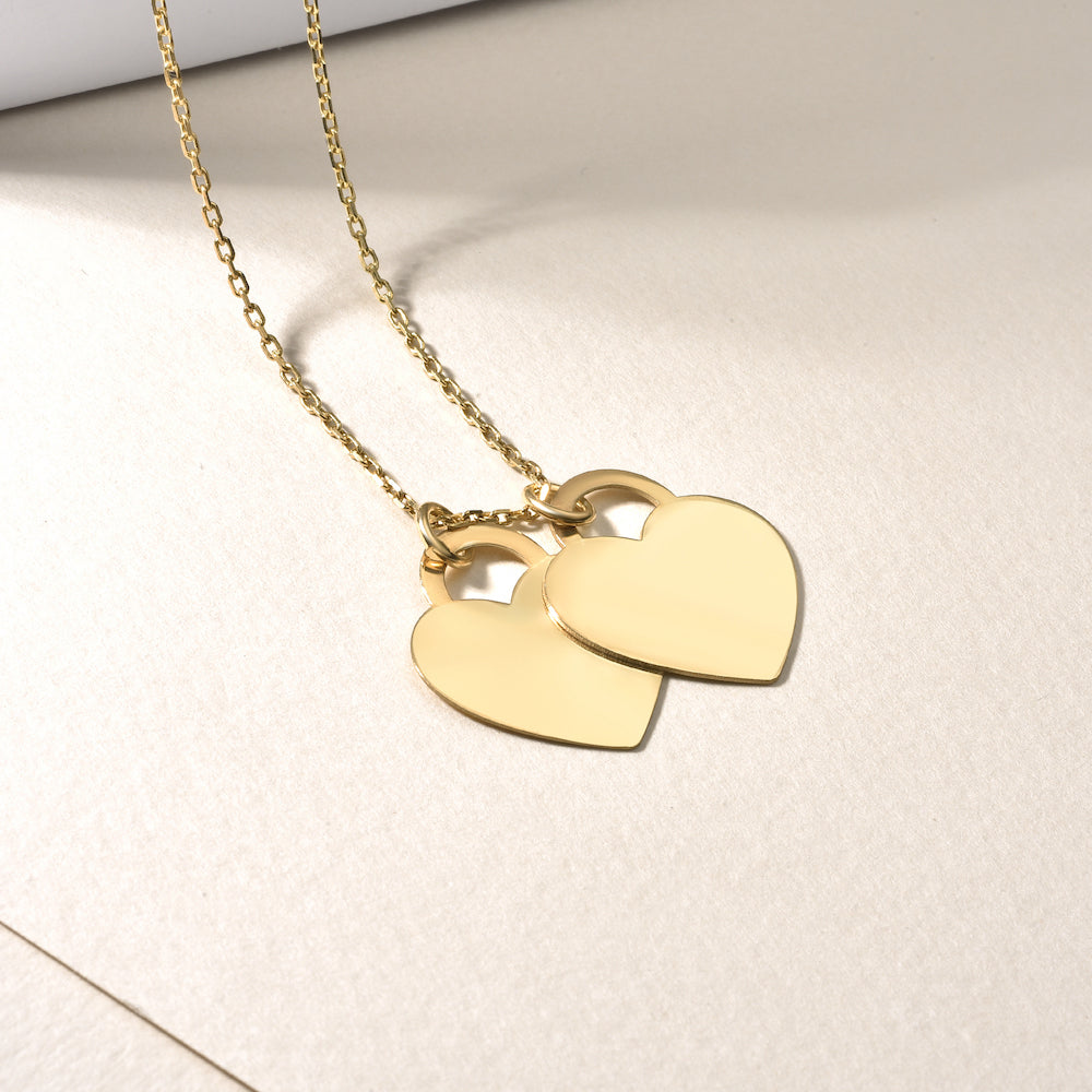 Double Heart Halskette in Gold auf hellem Stoff, zwei glänzende Herz-Anhänger an feiner Kette.