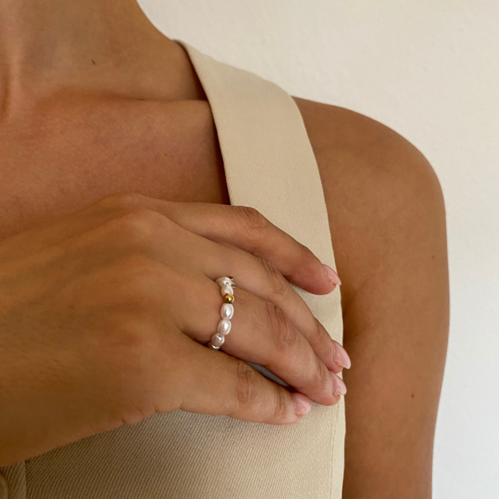 Damenhand mit Perl Ring aus weißen Perlen und goldener Kugel, cremefarbenes Top.