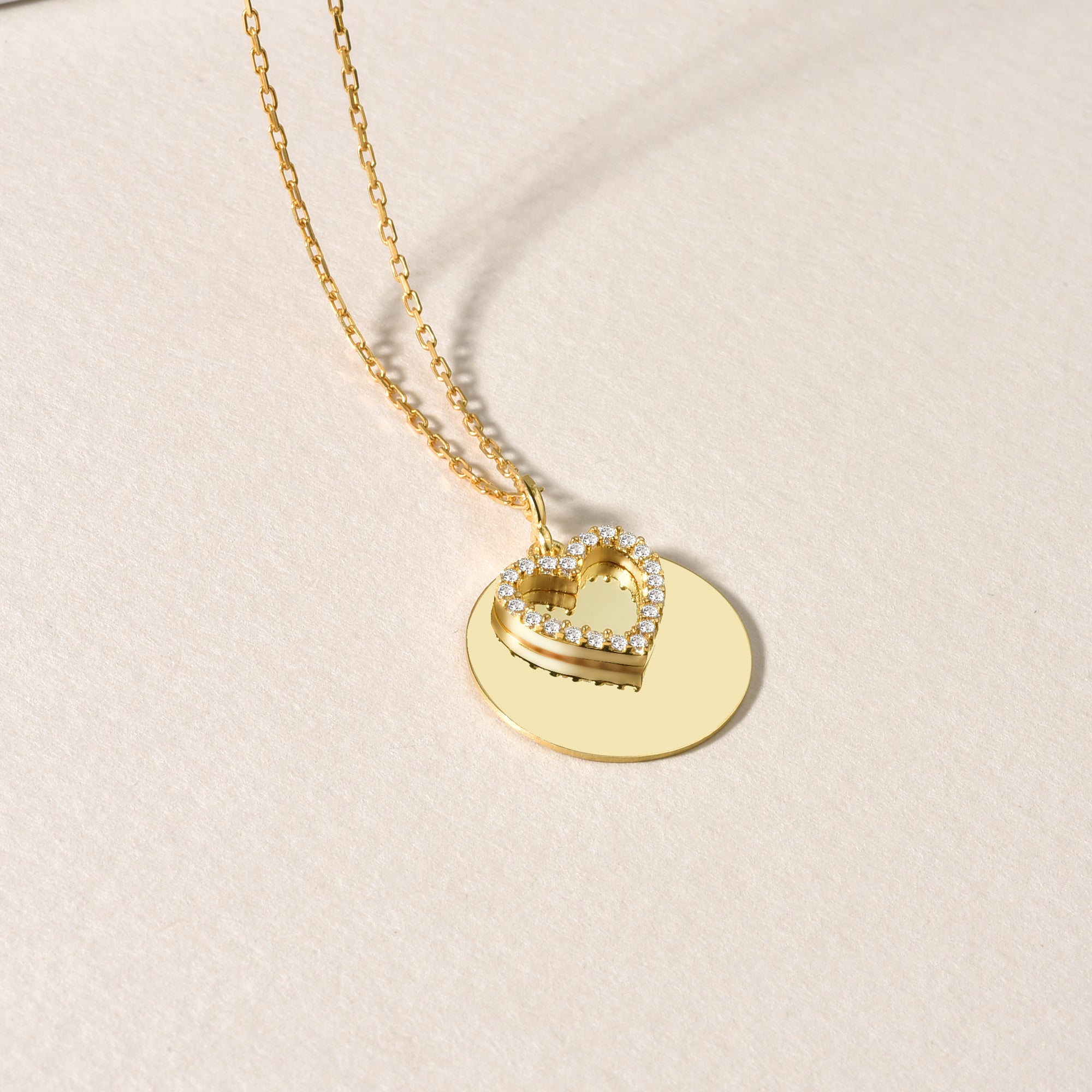 Goldene Halskette mit Herz-Zirkonia und rundem Anhänger, seitliche Detailaufnahme auf hellem Untergrund.