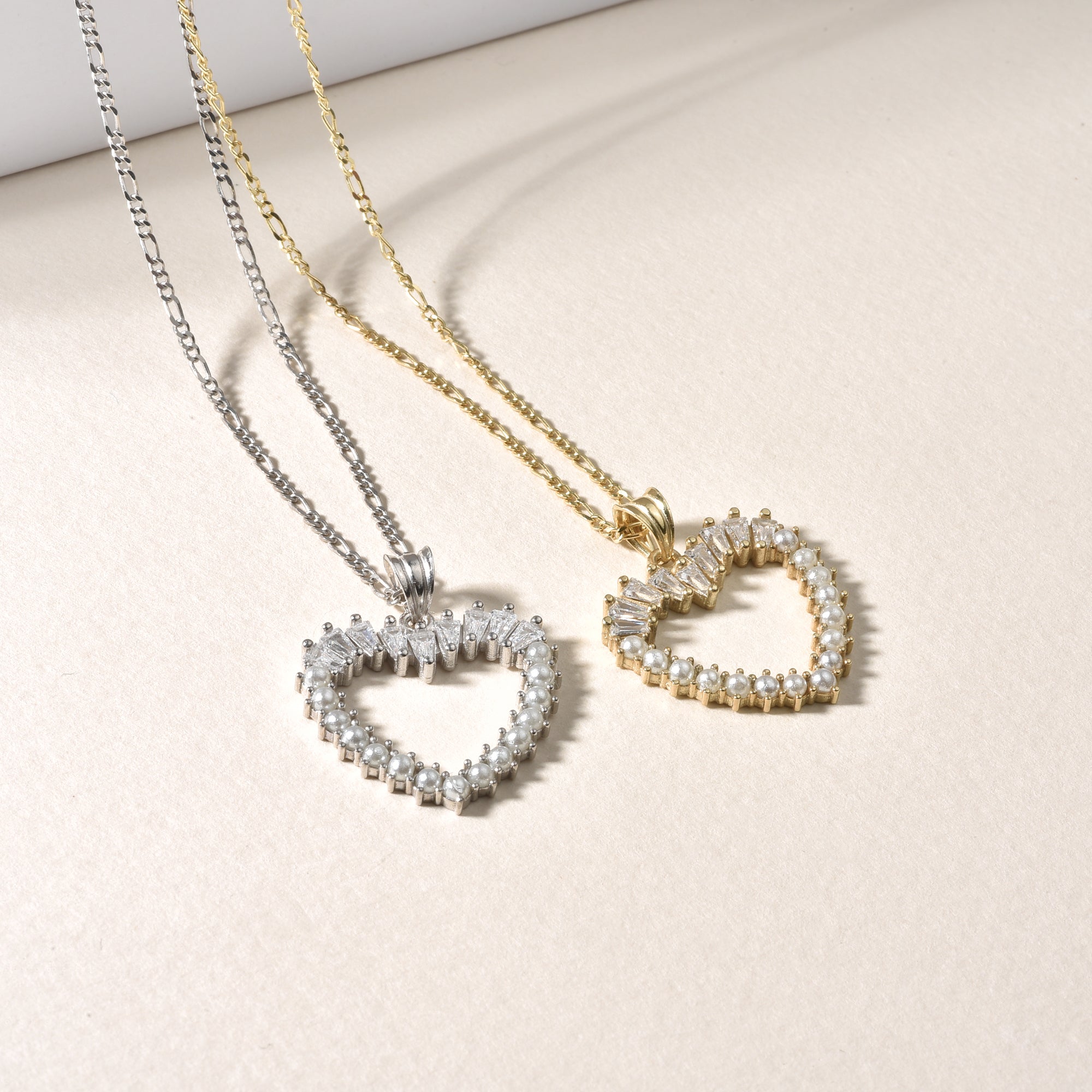 Vergleich Gold & Silber: Herz-Halsketten mit Perlen und Baguette-Zirkonia auf hellem Untergrund.