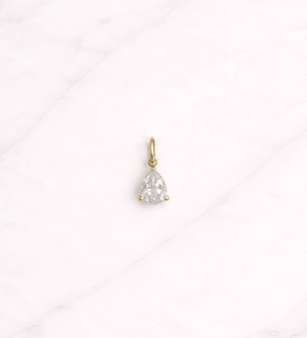 Manhattan CHARM in Gold mit tropfenförmigem klarem Stein auf hellem Marmorghintergrund.