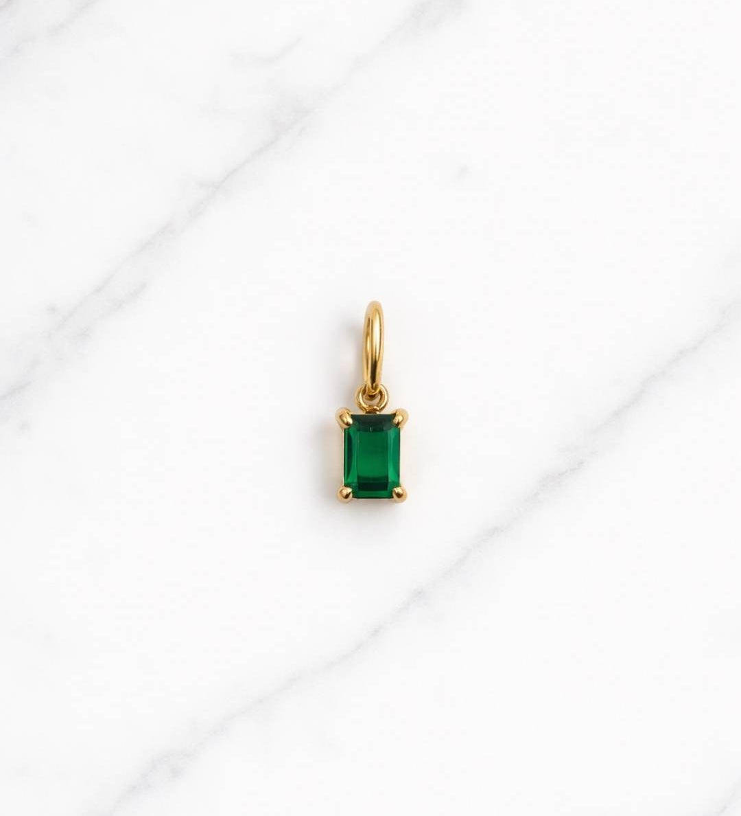 Monte Carlo Charm in Gold mit rechteckigem grünem Stein auf hellem Marmorhintergrund.