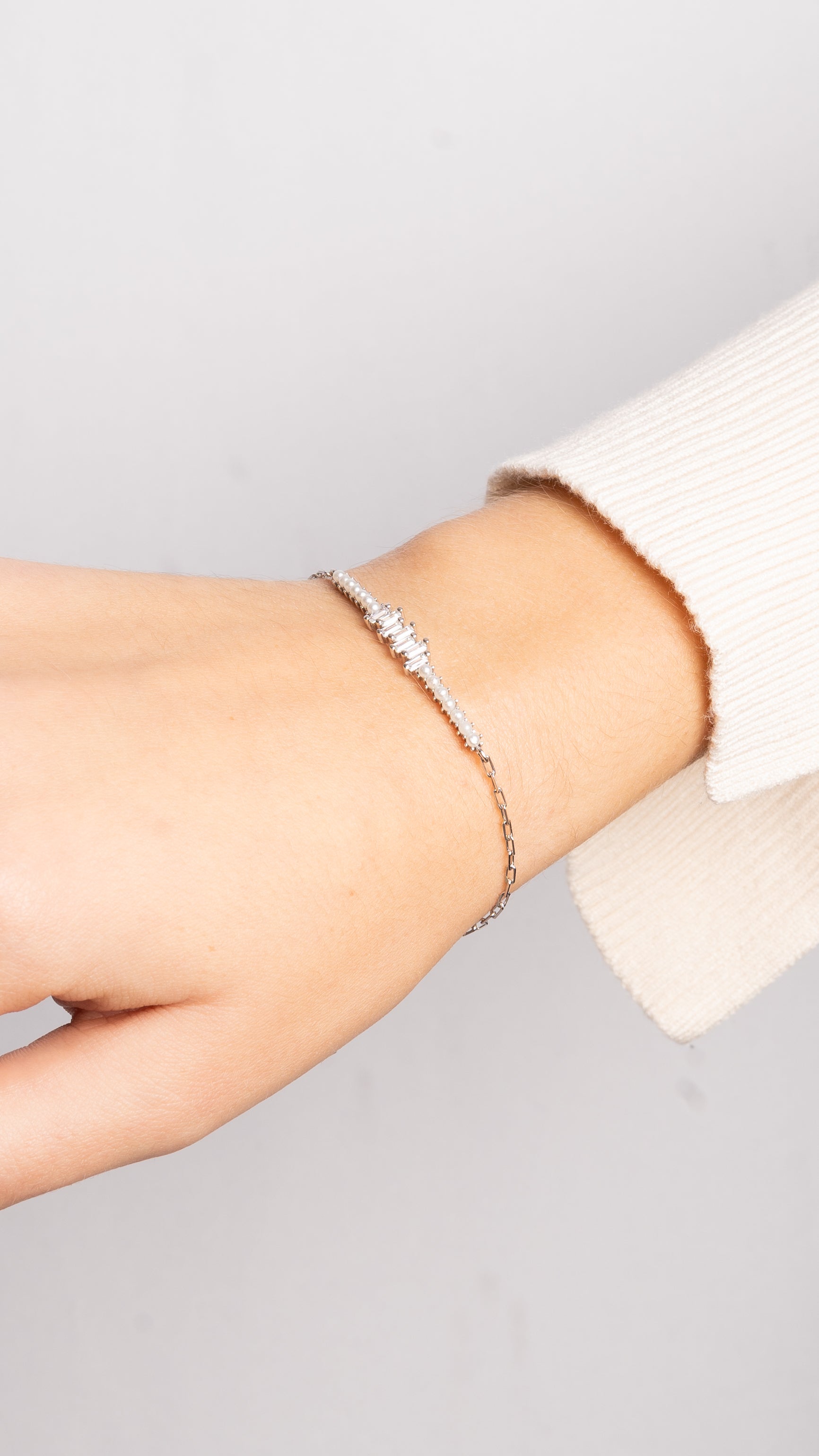 Zarte Pearl X Baguette Armkette in Silber an Handgelenk mit cremefarbenem Pullover.