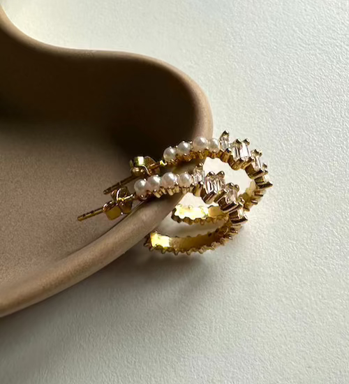 Goldene Pearl X Baguette Ohrringe mit Perlen und Zirkonia, liegend auf Keramik-Schale.