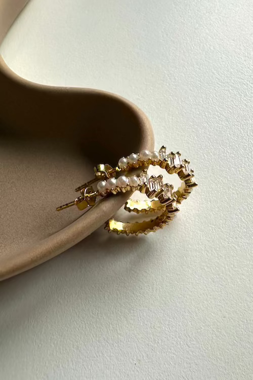 Goldene Pearl X Baguette Ohrringe mit Perlen und Zirkonia, liegend auf Keramik-Schale.