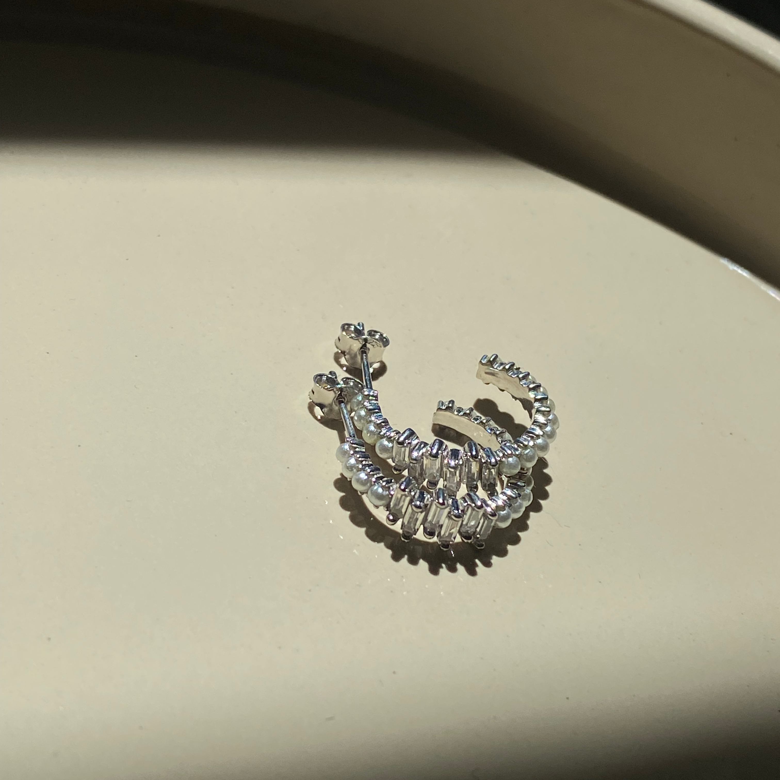 Pearl X Baguette Ohrringe in Silber mit Perlen und Zirkonia auf beigem Untergrund.