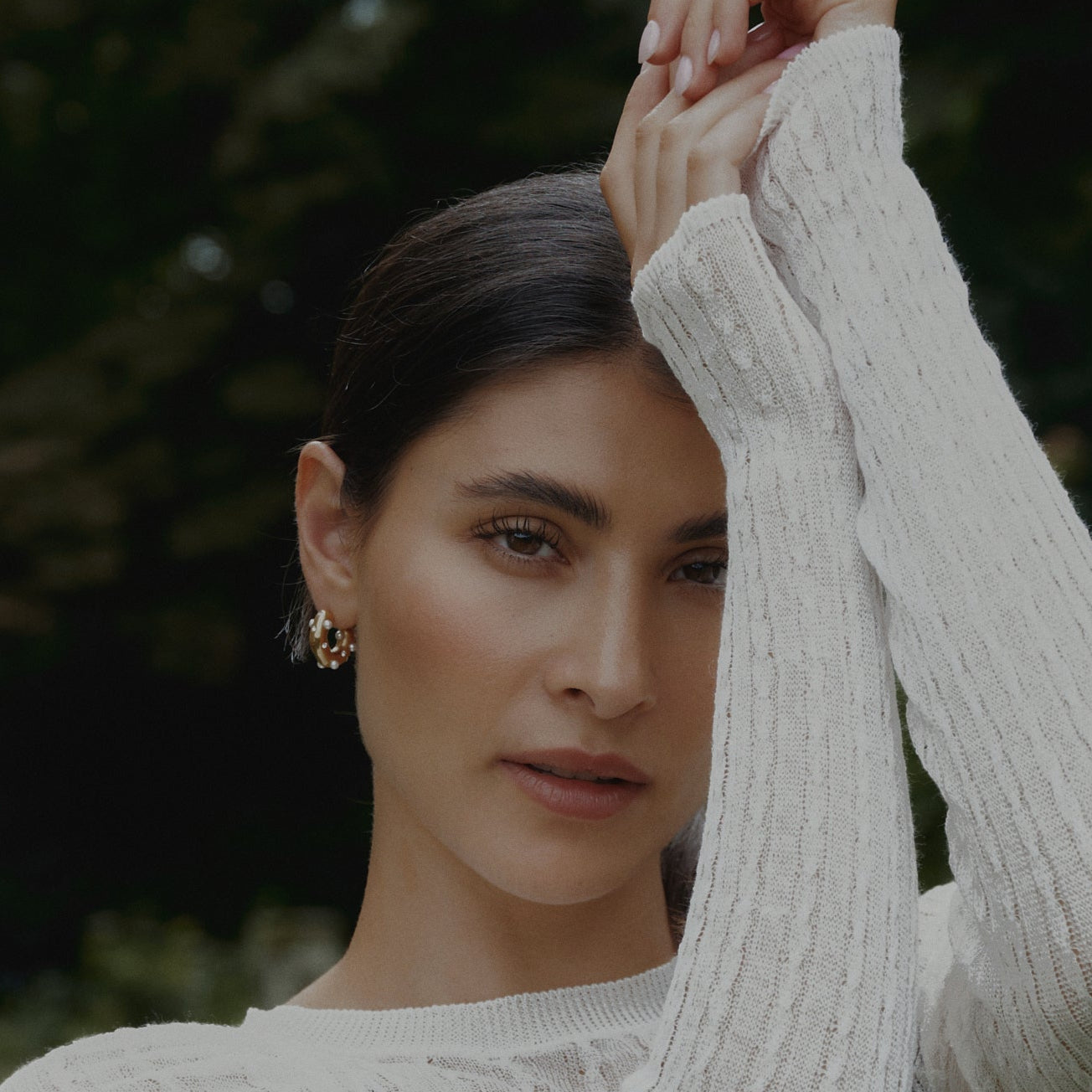 Model im weißen Strickpullover mit Perl Zirkonia Creolen Gold vor dunklem Grün.