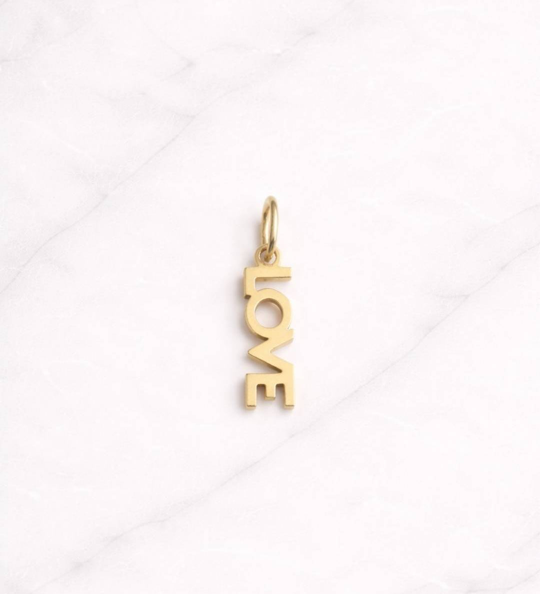 Positano CHARM in Gold mit vertikalem Schriftzug auf hellem Marmorhintergrund.