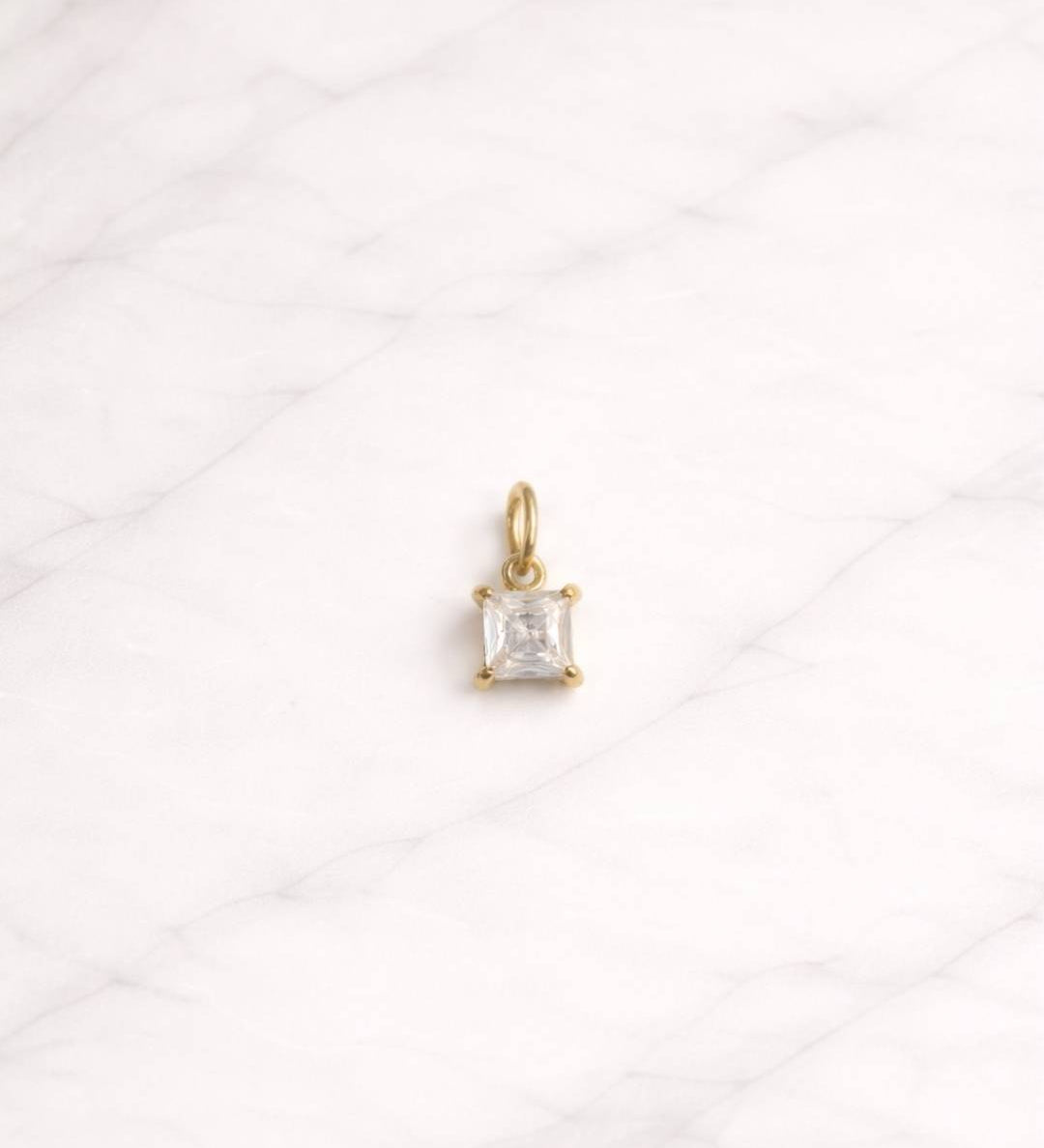 Singapore CHARM in Gold mit quadratischem klarem Stein auf hellem Marmorhintergrund.