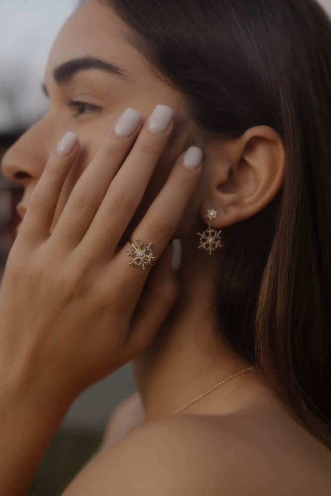 Frau trägt Snowflake Ice X Ring Gold am Finger, funkelndes Schneeflocken-Design passend zu Ohrringen.