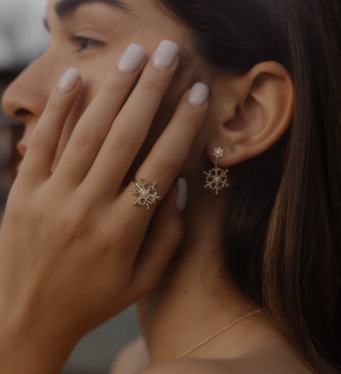Frau trägt Snowflake Ice X Ring Gold am Finger, funkelndes Schneeflocken-Design passend zu Ohrringen.