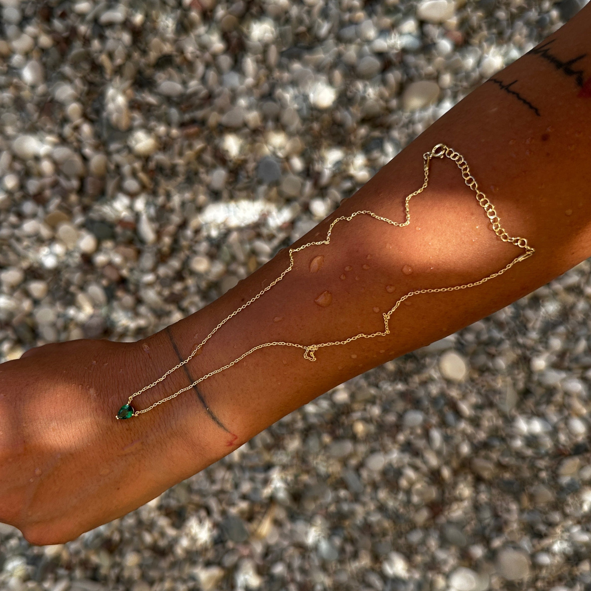 Goldene Solitär Drop Green Chain auf Arm im Sonnenlicht, grüner Tropfenanhänger sichtbar.
