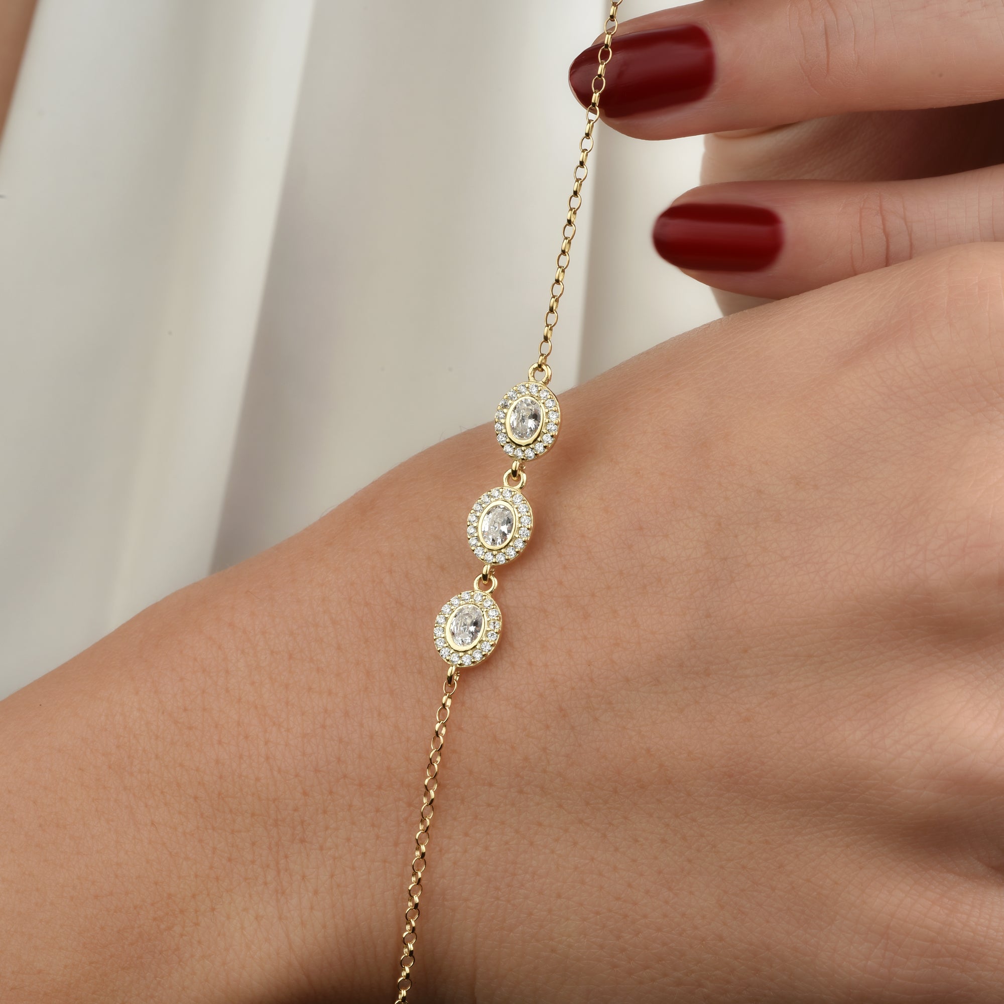 Hand hält das goldene Solitaire Kranz Oval Armband, Detail der drei ovalen Zirkonia mit Halo-Fassung.