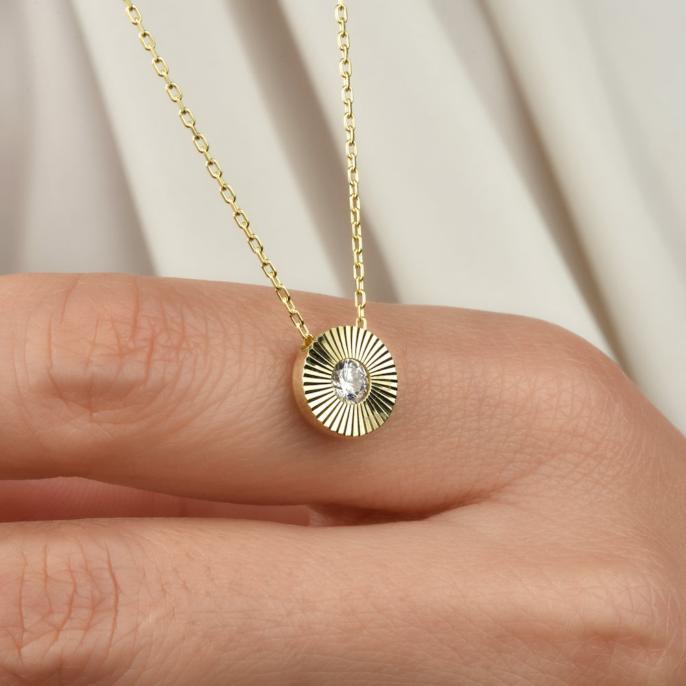 Goldene Solitaire Sun Halskette mit Sonnenstrahlen-Anhänger und Zirkonia auf einem Finger vor hellem Stoff.
