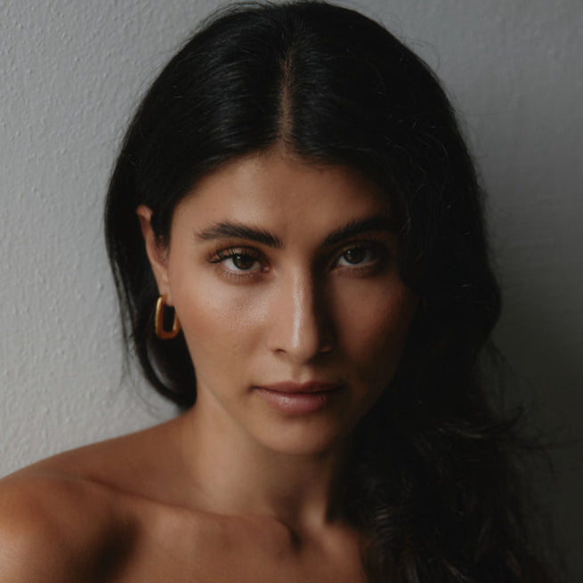 Frau mit dunklem Haar trägt einen goldenen Square Hoop Ohrring, eleganter Portrait-Look.