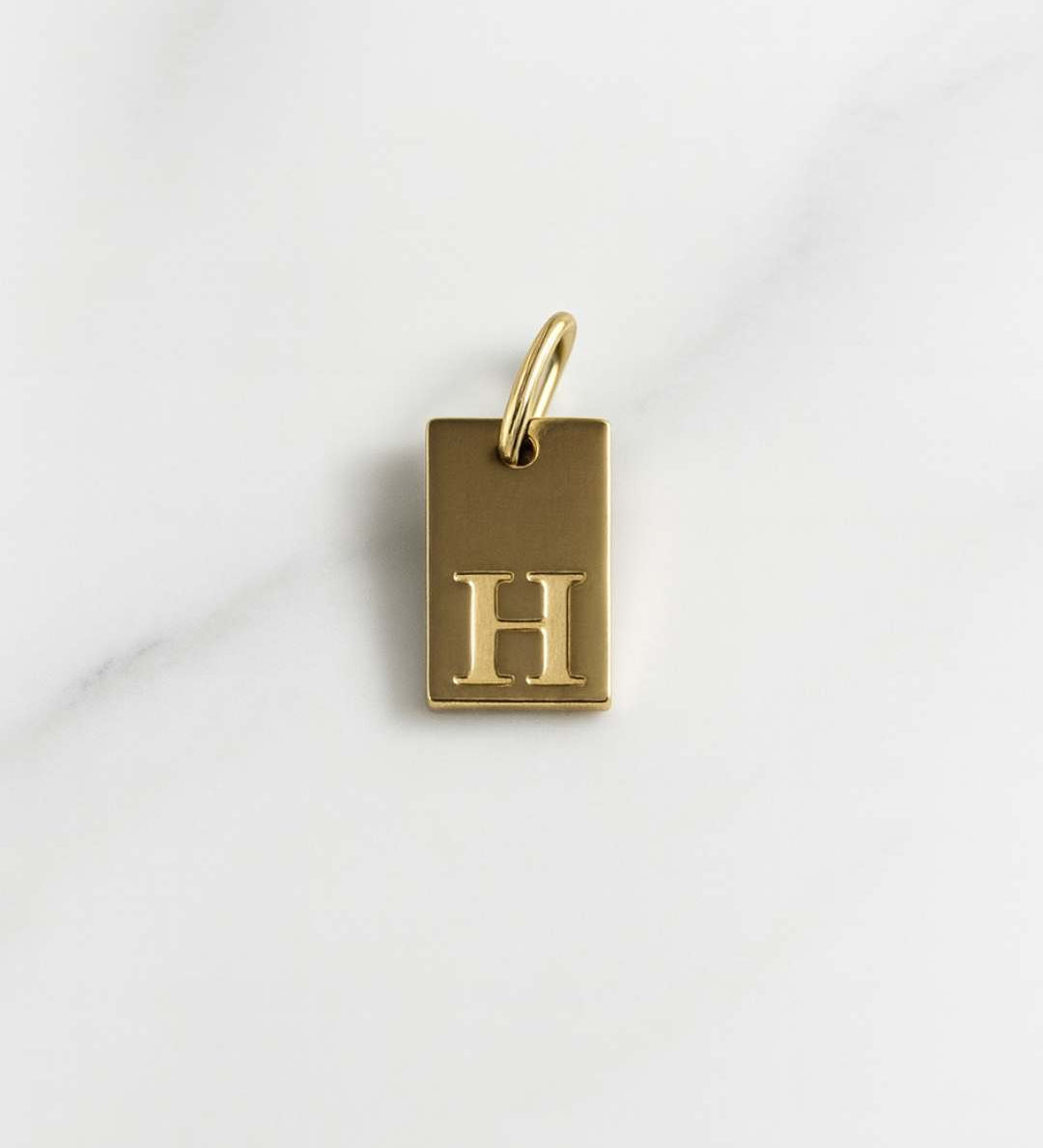 Vergoldeter rechteckiger Initial Charm der Tokio Kollektion auf hellem Marmorfond