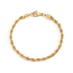 Goldenes Twist Armband aus Edelstahl neben flacher Kette auf hellem Untergrund.