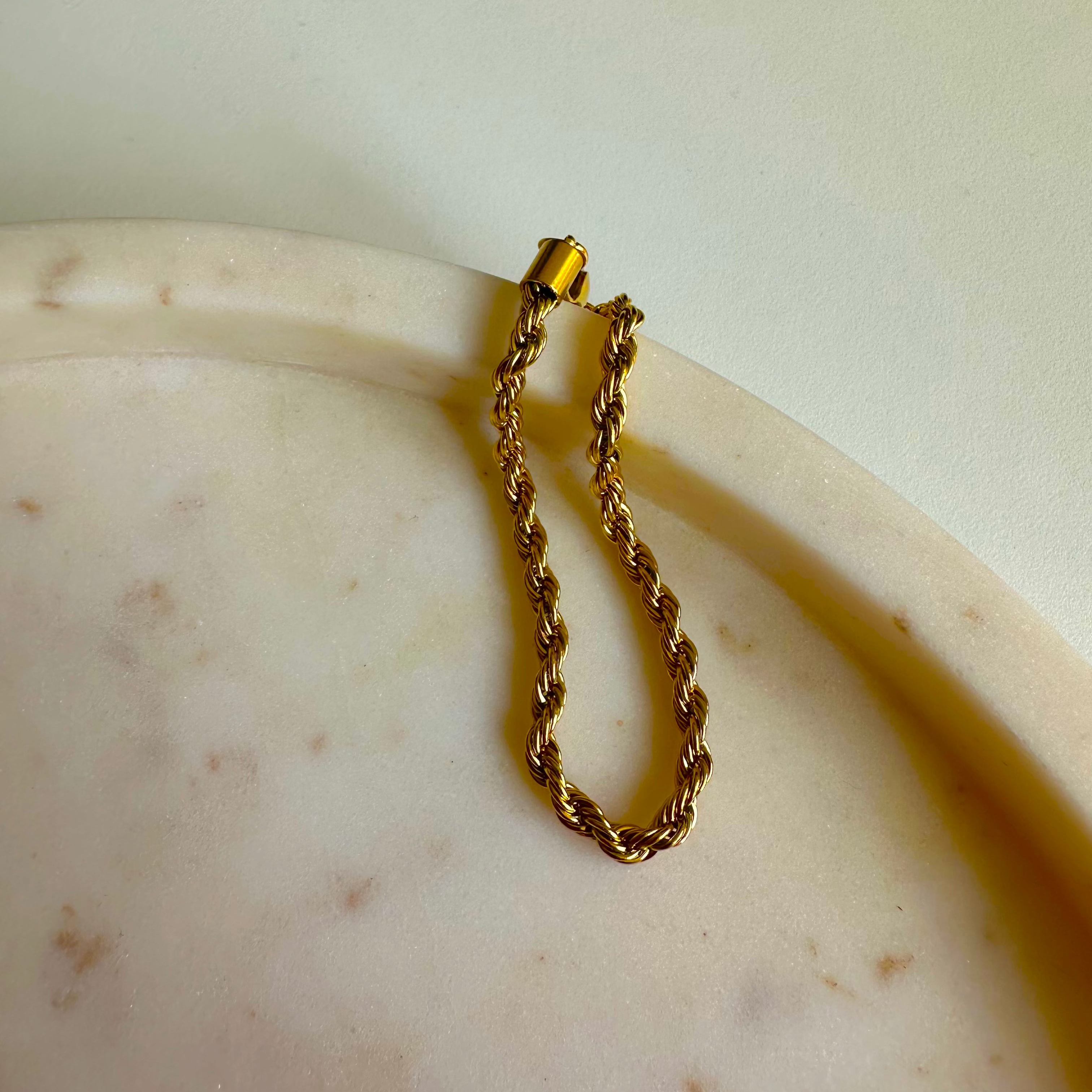 Goldenes Twist Armband aus Edelstahl auf heller Steinplatte dekorativ gelegt.