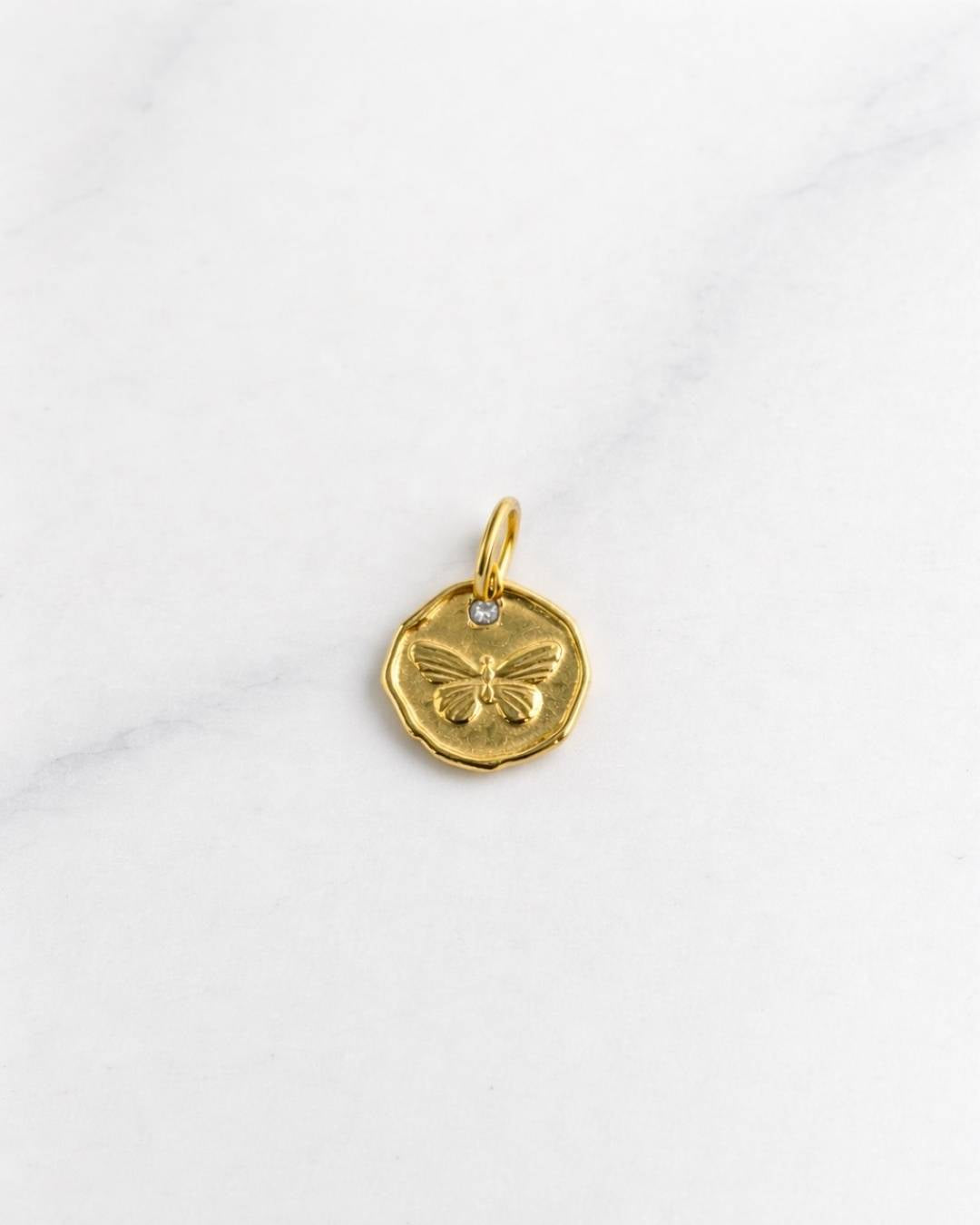 Vienna Charm in Gold mit Schmetterling-Prägung und kleinem Stein auf hellem Hintergrund.