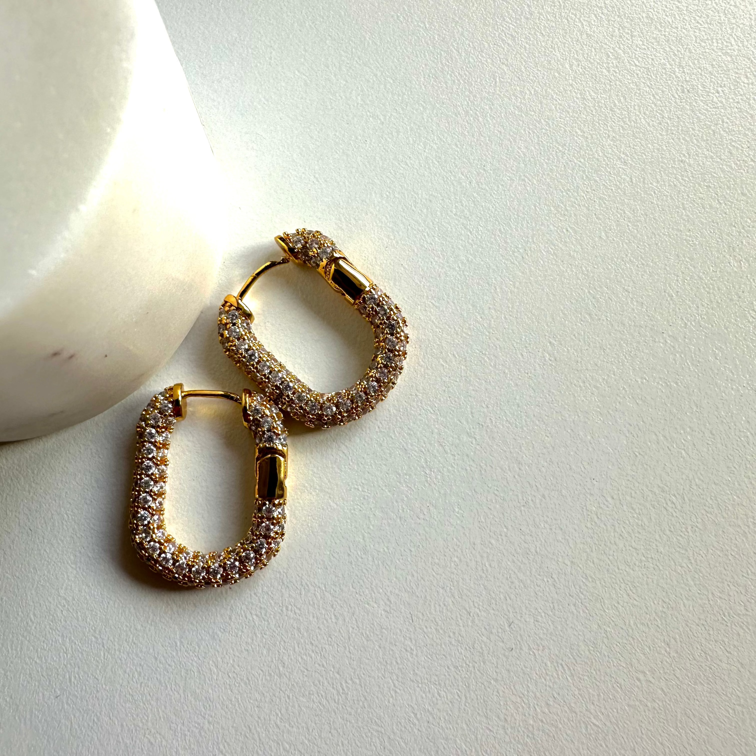 Zwei goldene eckige Zirkonia Hoop Ohrringe liegen dekorativ auf hellem Untergrund.